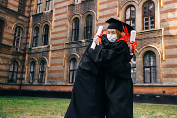 Fototapeta na wymiar Graduates in dressing gowns with diplomas in their hands stand next to the university and hug. Wearing protective masks against disease.