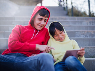 two brothers play a video game with a tablet in the park. the older brother teaches the younger brother.