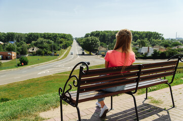  A young girl sits on a bench and looks towards the leaving road. Place for your text.