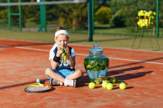 A Small Kid In A Tennis Suit Sitting On A Tennis Court In The Summer And Drinking Cold Lemonade. Dispenser With A Fresh Drink And Tennis Balls In The Background.