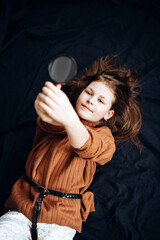 Cute girl looks at a magnifying glass while lying on a dark background