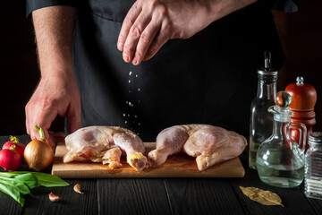 The cook adds salt to the raw chicken legs. Preparation for grilling with chicken leg in the kitchen of a restaurant or cafe on table with vegetables and spices