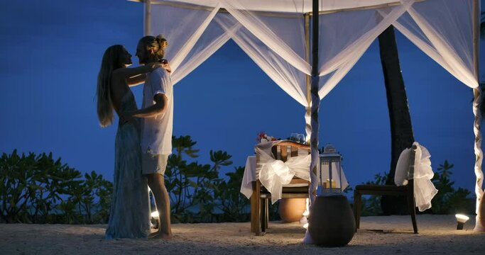Young lovely couple dancing and kissing beside a dinner table on a tropical beach with magic moment celebrating anniversary relationship. Happy husband and wife have romantic time in valentines day. 