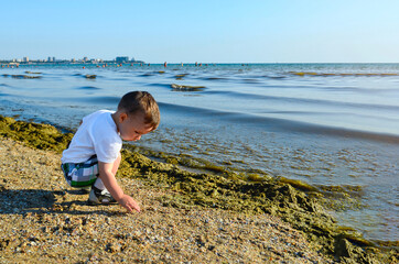 Emotional portrait of a child brightly lit. Looks concentrated, squints from the sun. Wearing a hat against the blue sea and sky, on the shore on a hot summer day.