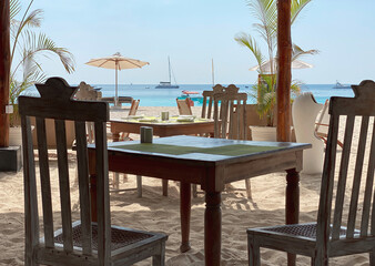 Restaurant tables against the backdrop of the ocean, ships, palm trees and beautiful white sand. Beach vacation and travel concept.