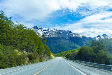 Fototapeta premium estrada de asfalto rodeada de vegetação verde típica da patagônia e ao fundo montanhas com neve e um belo céu azul.