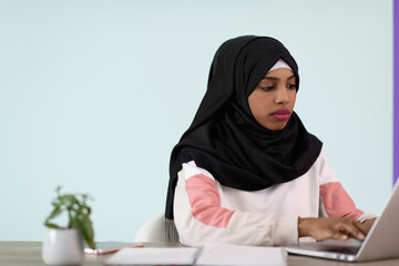 afro girl wearing a hijab thoughtfully sits in her home office and uses a laptop
