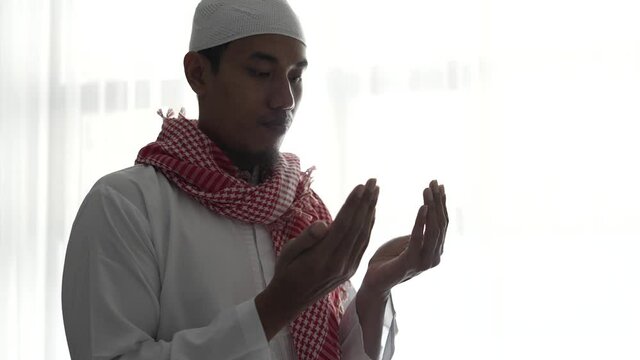 portrait of muslim man using hand to pray against white window