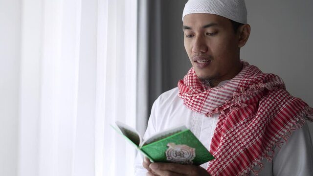 asian religious man reading koran or quran while standing next to the window