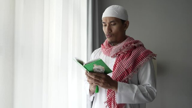 asian religious man reading koran or quran while standing next to the window