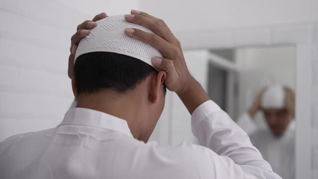 muslim asian man looking at mirror and get dressed before going to the mosque wearing islamic hat or cap