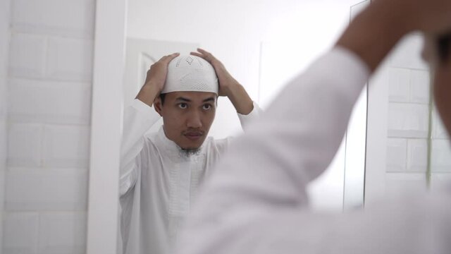 muslim asian man looking at mirror and get dressed before going to the mosque wearing islamic hat or cap