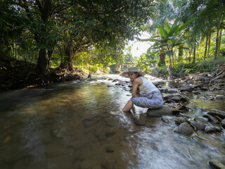 Young Asian woman with straw hat sitting on the rock and enjoying waterfall landscape. Travel lifestyle. 