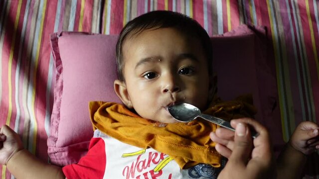 A mother feeds her 7-month-old baby soft food and water using a steel spoon. An infant baby is eating to lying down. Baby looking at camera. Top view. Slow-motion video. 