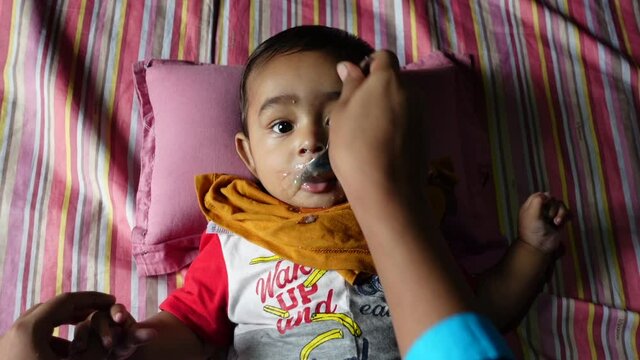 A mother feeds her 7-month-old baby soft food and water using a steel spoon. An infant baby is eating to lying down. Baby looking at camera. Top view. Slow-motion video. 