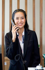 Asian hotel receptionist in black suit answer the phone while smiling