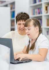 Young girl with down syndrome  uses a laptop with her teacher at library. Education for disabled children concept