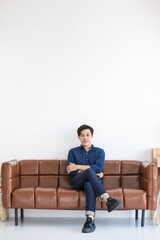 Young handsome man in blue shirt and jeans is sitting with his leg crossed at the middle on a big dark brown sofa with smile.