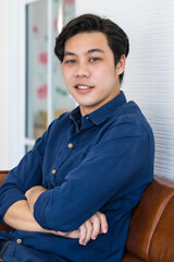 Fototapeta premium A young man in blue shirt and blue jeans is sitting on a dark brown sofa with smile. He folds his arms in relax gesture