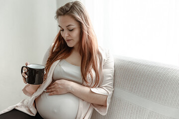 Young pregnant woman is resting on the sofa with a cup of hot drink copy space.