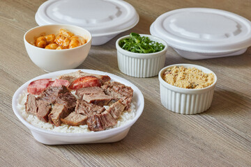 Marmitex with white rice, beans and barbecue with side dish of cabbage, crumbs and gnocchi with white packaging in the background on a textured wooden table.