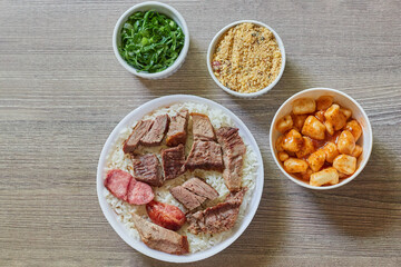 Marmitex with white rice, beans and barbecue with side dish of cabbage, crumbs and gnocchi on a textured wooden table - top view.