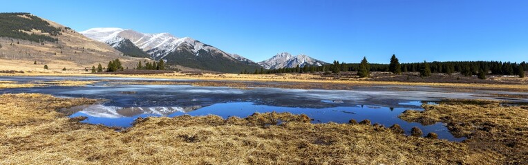 Naklejka na ściany i meble Scenic Panoramic Landscape View of Prairie Grassland and Distant Mountain Peaks Skyline reflected in Calm Blue Lake Water. Alberta Foothills of Canadian Rockies on a sunny Springtime Day