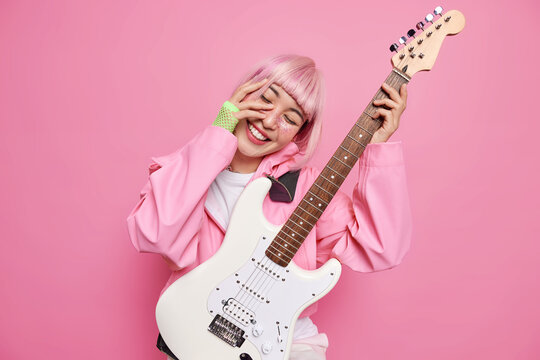 Celebrities concept. Positive stylish female guitarist tilts head smiles happily keeps hand on face performs rock music on white acoustic guitar isolated over pink wall. Woman hols musical instrument
