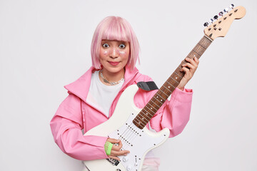 Fototapeta premium Indoor shot of popular female artist plays rock music on electric guitar poses against white background with musical instrument wears pink jacket has glitters on face. Popular singer has rehearsal