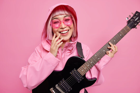 Pleased fashionable woman dressed in stylish jacket pink sunglasses has fun pretends to be rock star smiles broadly uses electric guitar isolated over rosy background. Talented musician indoor