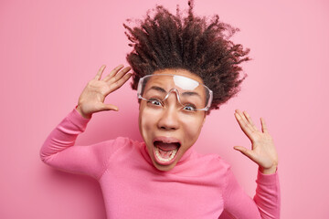 Emotional young Afro American woman screams loudly has funny face hair floating in air keeps hands raised wears transparent glasses turtleneck isolated over pink background feels super crazy