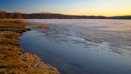 Colorful sunset on the shore of a lake in early spring. Reeds and trees in the distance. The surface half-covered with ice.