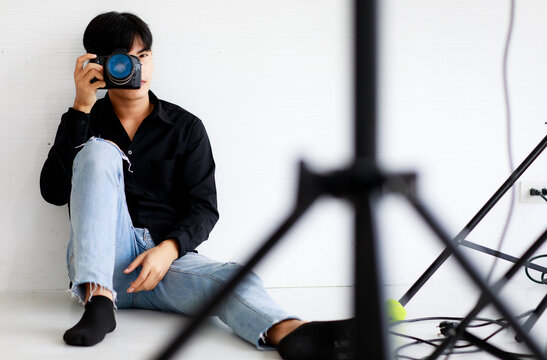 Asian handsome male model wearing casual black shirt with jeans, holding a camera, sitting on white background in a studio and posing while put his hand on hair.