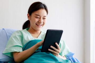 An asian young pretty female patient smiling with happiness and touching screen of tablet for online chat or communicate while lying on bed in hospital. Technology and Medical Concept.