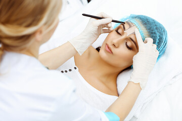 Naklejka premium Professional beautician is going to correct eyebrow line at woman face using cosmetic ruler. Permanent brow makeup in beauty salon, closeup. Cosmetology treatment