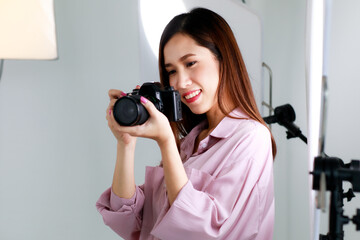 An asian beautiful adult female photographer with long hair wearing casual shirt, holding a camera and taking photo in indoor studio with blur background of  lighting equipments.