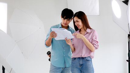 Portrait of two handsome man and beautiful woman wearing casual shirts holding paper of  script or picture reference while standing in indoor photo studio with blur background of lighting equipments.