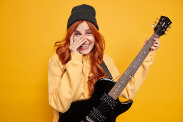 Happy female guitar player with natural red hair chuckles and keeps hand on face plays favorite...