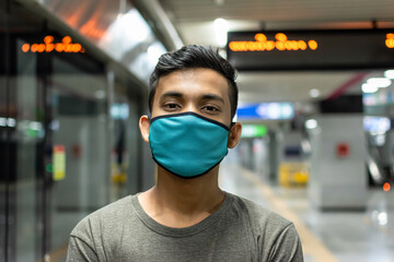 Portrait of handsome young man with a medical mask in casual style t-shirt standing, raised arms and looking at camera with smile in a metro station.