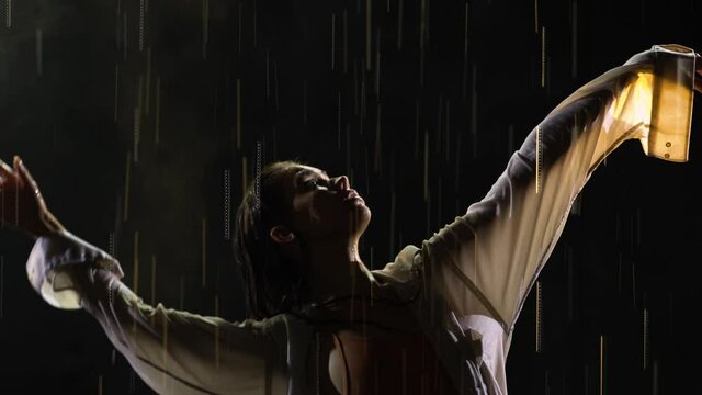 Wet young woman in white shirt and beige bodysuit is dancing dramatic modern ballet dance in rain. Ballet dancer moves plastic, performing modern choreography on surface water in dark studio. Close up