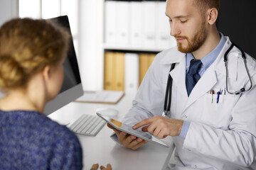 Friendly red-bearded doctor and patient woman discussing current health examination while sitting and using tablet computer in clinic
