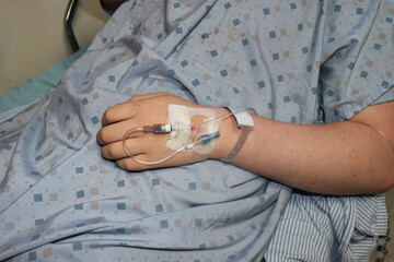 Person on a hospital bed, wearing a gown with an IV in the back of their hand.
