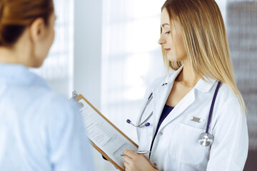 Young woman-doctor and her patient are discussing patient's current health examination, while...
