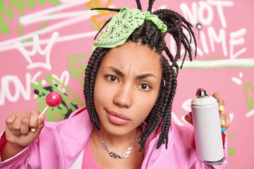 Close up shot of beautiful Afro American teenage girl looks attentively at camera holds lollipop and aerosol spray creats colorful graffiti wall in urban place being street vandal or hooligan