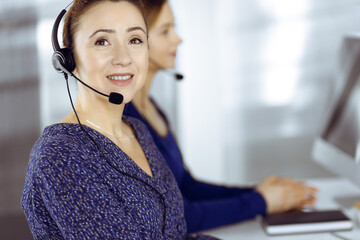 Two busineswomen have conversations with the clients by headsets, while sitting at the desk in a...