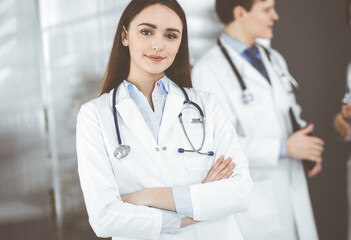 Smiling intelligent woman-doctor is standing with arms crossed in a clinic, together with her colleagues at the background. Portrait of physicians at work. Perfect medical service in a hospital