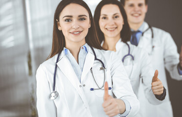 Group of young cheerful doctors is standing as a team with thumbs up in a hospital office and is ready to help patients. Medical help, insurance in health care, best disease treatment and medicine