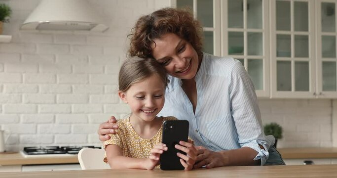 Young mom her little daughter spend time together in cozy kitchen having fun holding smartphone, enjoy video call event, taking selfie using photo editing app laughing, modern technology usage concept