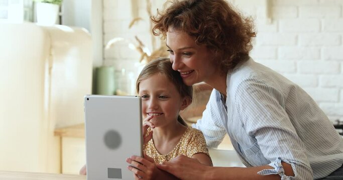 Young cheerful mother little 8s adorable daughter sit in kitchen at home spend free time with tablet device. Having fun on internet, studying online. Children safe usage of modern technology concept