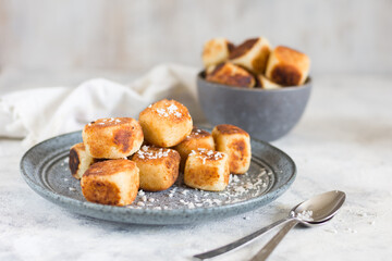 Square curd pancakes, traditional Russian cheese cakes in a gray plate and bowl, with coconut flakes. On a light background with instruments.